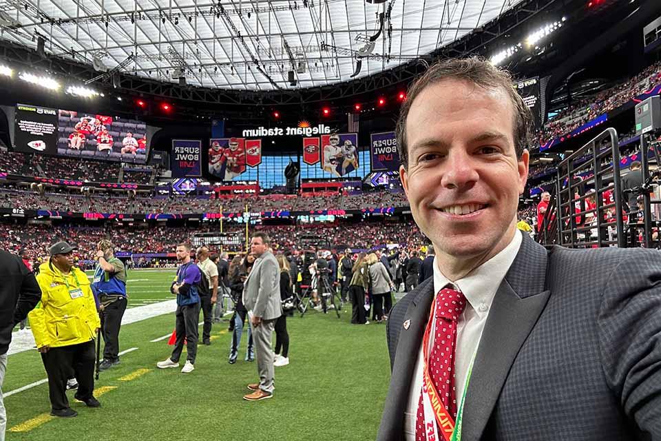 A man wearing a suit and a lanyard that says chiefs poses for a photo on a football field with a full stadium behind him. Signage reading Allegiant Stadium and Super Bowl are visible.